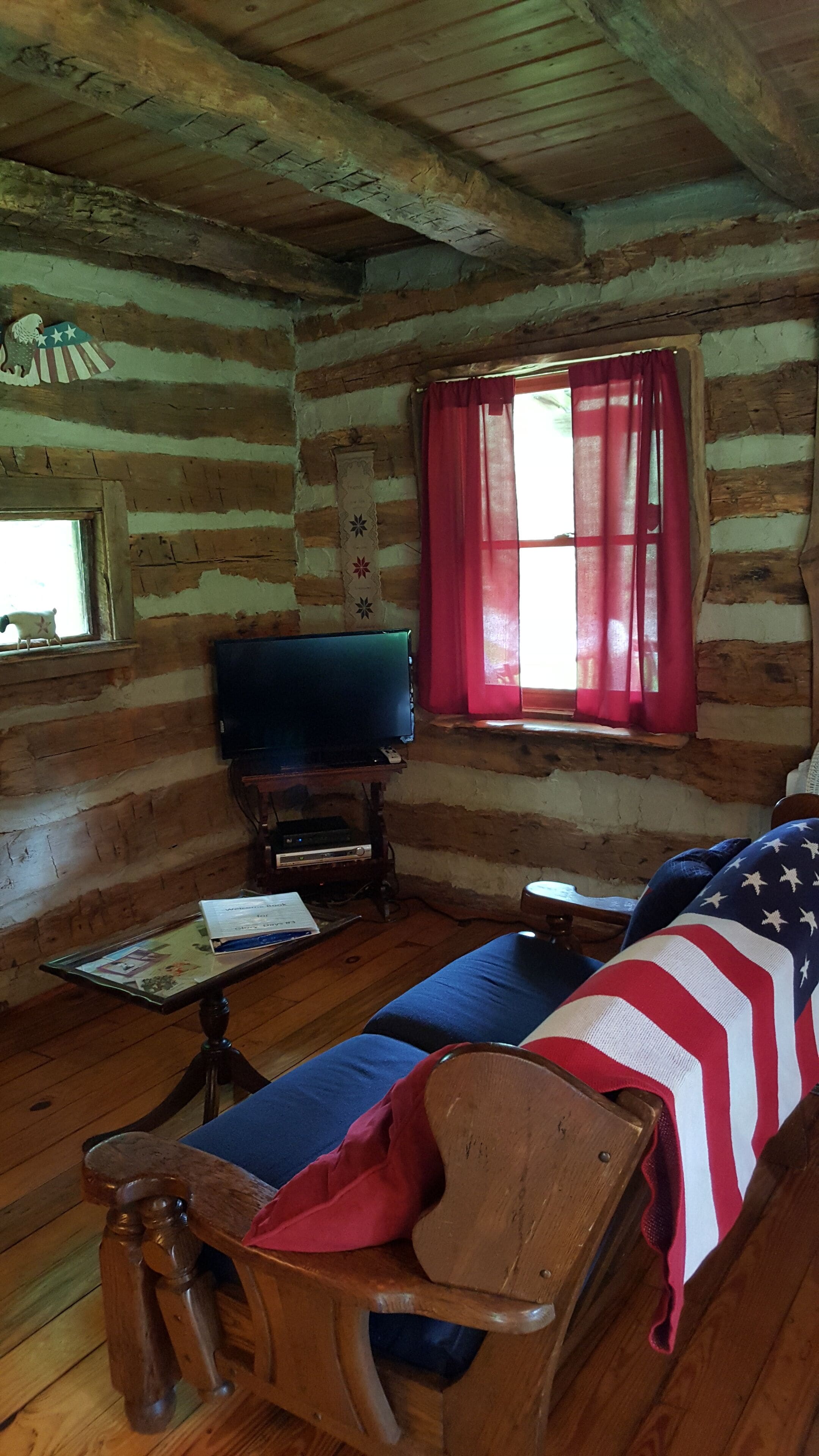A log cabin interior with a wooden sofa, a television, and a window with red curtains.