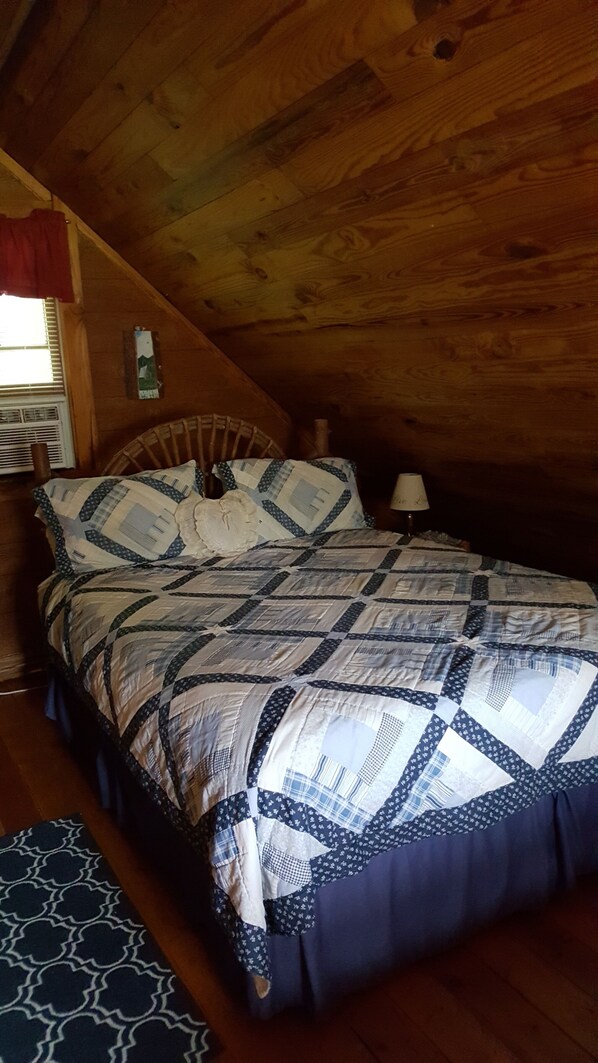 A bedroom with a wooden ceiling, a bed with a quilt, a nightstand, and a lamp.
