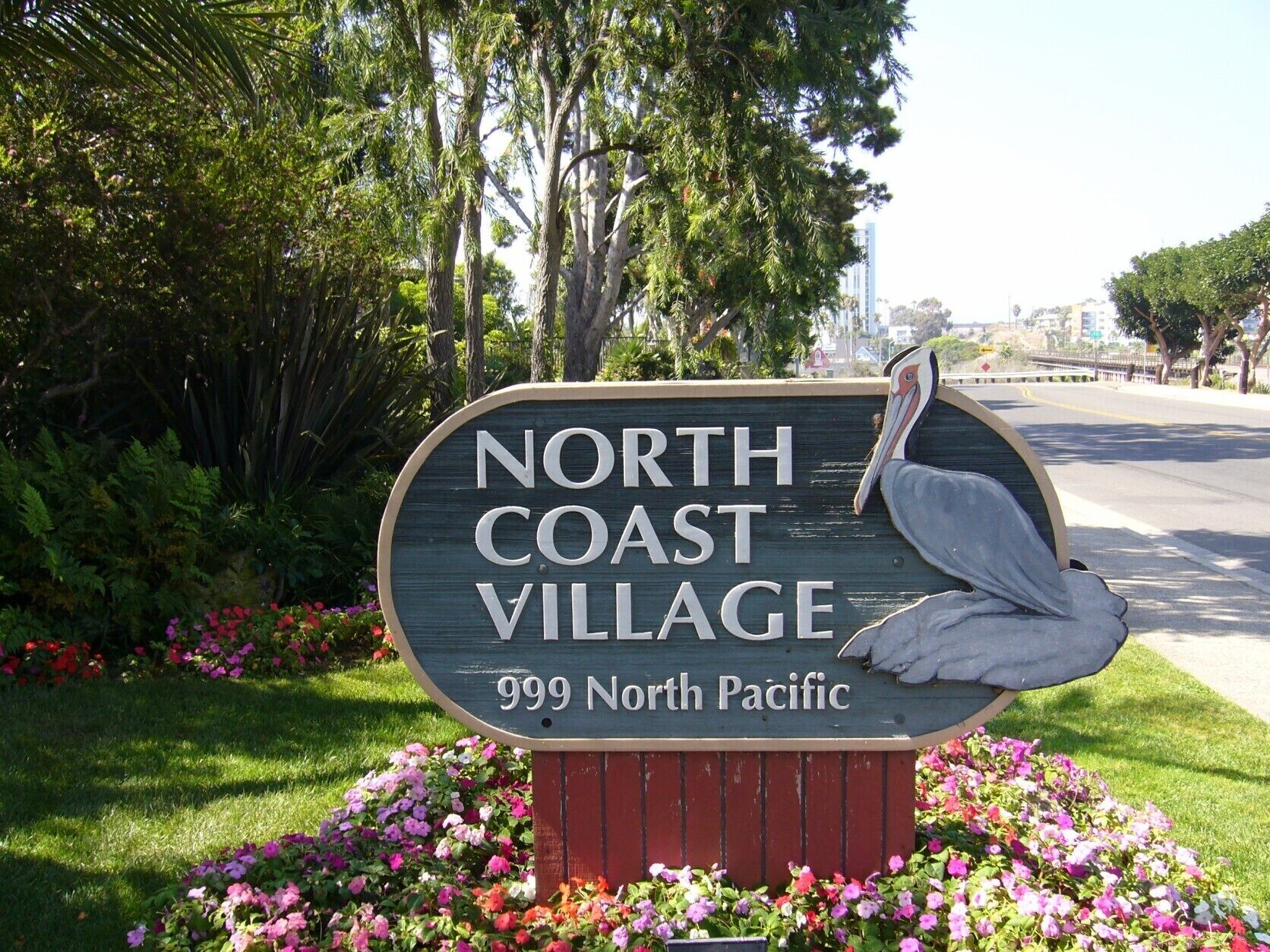 A sign for North Coast Village with a pelican sculpture, surrounded by colorful flowers.