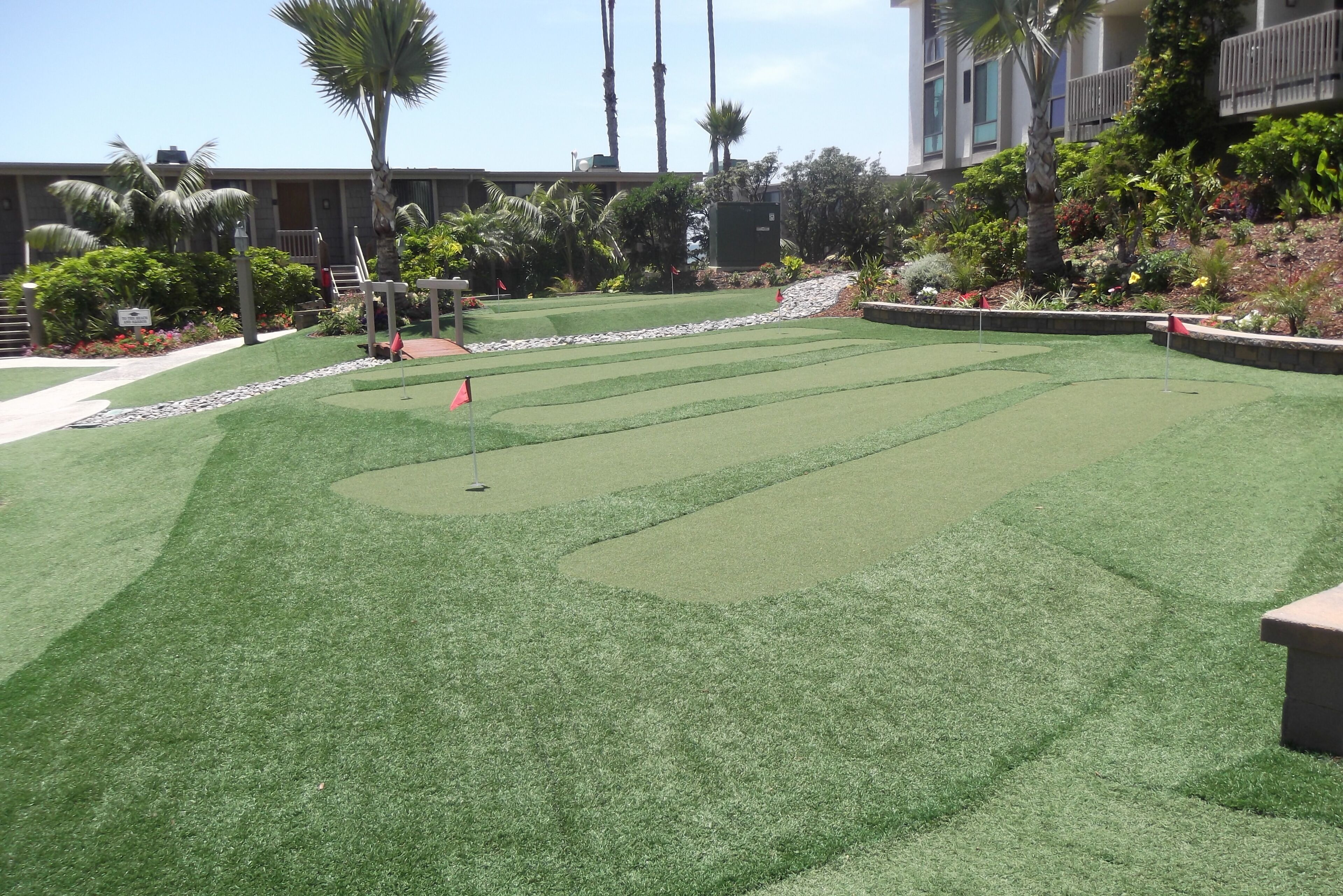A well-maintained artificial grass golf course with a putting green, surrounded by landscaped areas and palm trees.