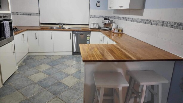 A compact kitchen with white cabinets, a stainless steel sink, a dishwasher, and a coffee maker. There are two stools and a small table.