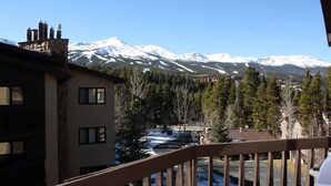 Une vue sur une montagne, avec des sommets enneigés, une rivière et un bâtiment doté d’une façade en pierre et de nombreuses fenêtres.
