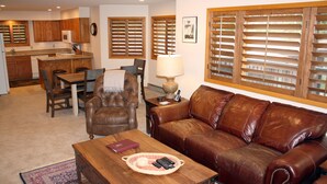 Un salon avec un canapé en cuir marron, une table basse en bois avec des télécommandes, un coin repas avec une table et des chaises, et une cuisine en arrière-plan.