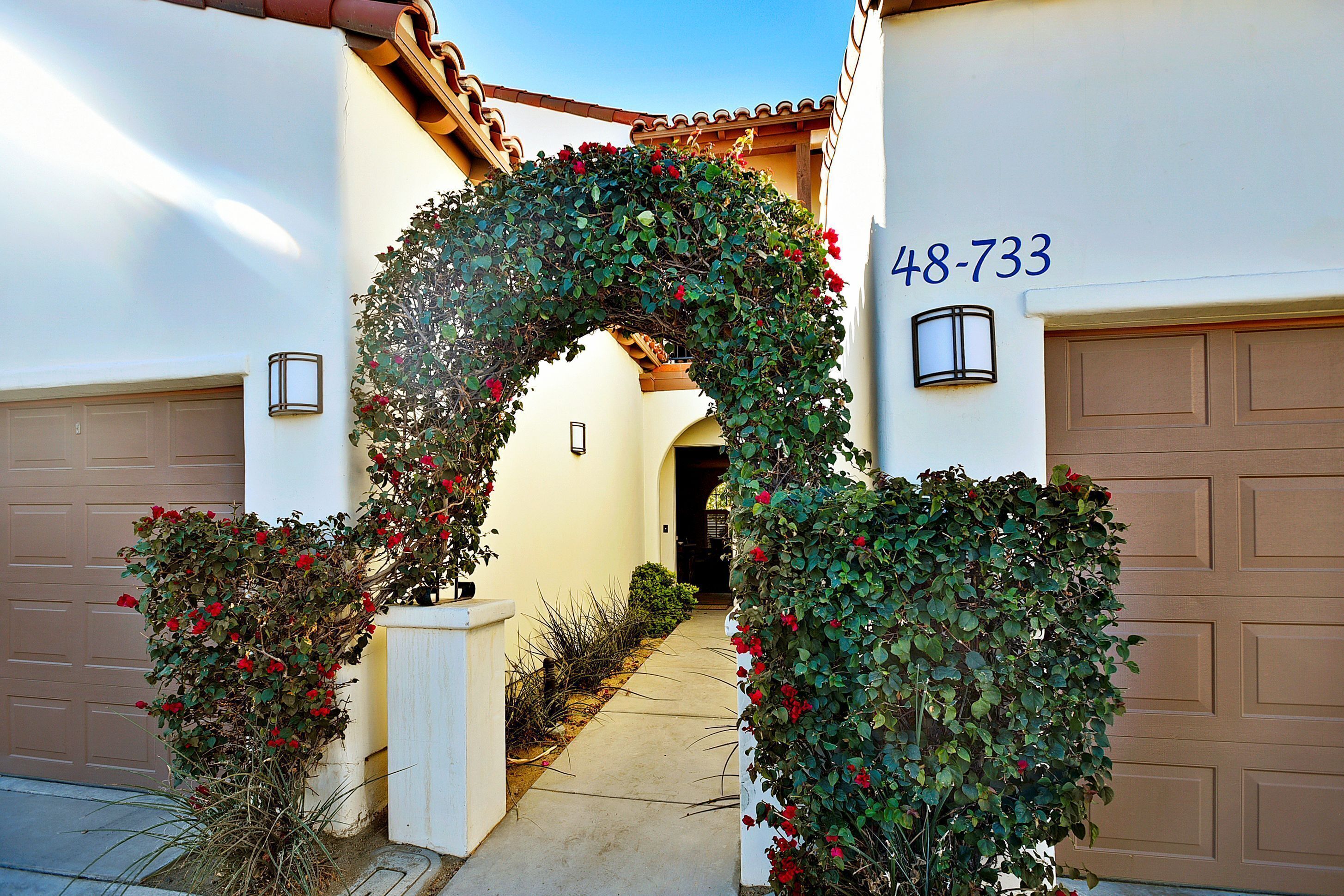 Une allée menant à une résidence, agrémentée d’une arche décorative recouverte de feuillage et de fleurs rouges, et aboutissant à un garage portant le numéro 48-733.