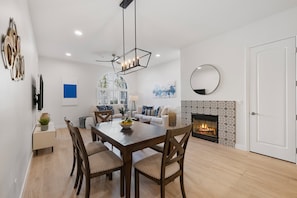 A modern living room with a fireplace, a dining area with a wooden table and chairs, and a large window providing natural light.