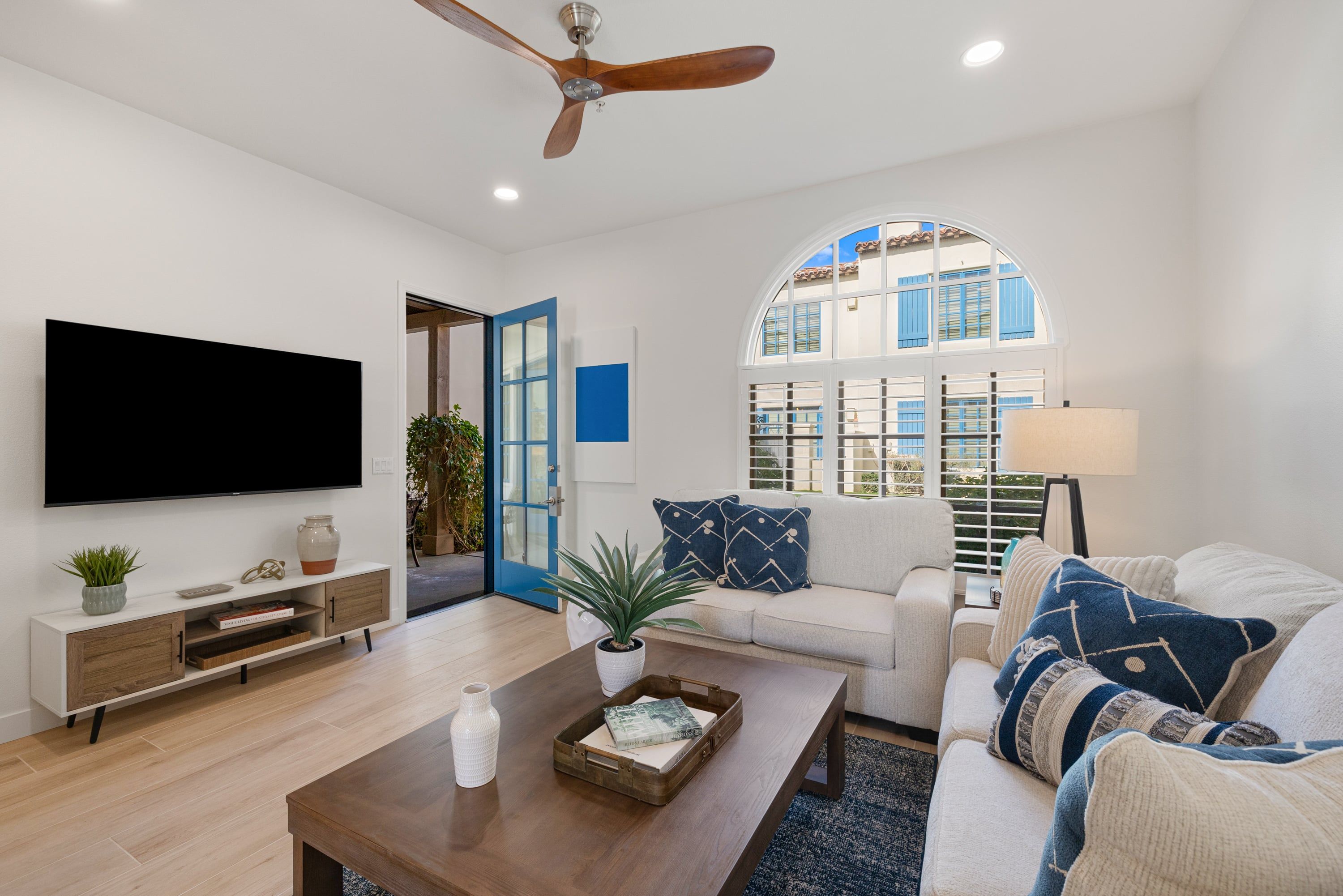 A modern living room with a flat-screen TV, a ceiling fan, a large window with a view, and a cozy seating area.