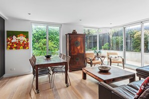 Ein geräumiges Zimmer mit Esstisch, Stühlen, einer Kommode und einer Couch. Große Fenster bieten Blick ins Grüne.