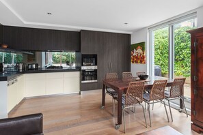 A modern kitchen with a dining area, large windows, and a view of greenery. - Lalique Elwood Beachside Apartment (Elwood)