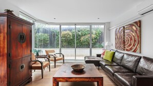 A modern living room with a wooden cabinet, leather sofa, and a large window offering a view of greenery. - Lalique Elwood Beachside Apartment (Elwood)
