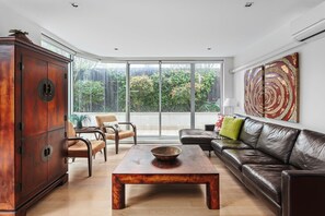 A modern living room with a wooden cabinet, leather sofa, and a large window offering a view of greenery. - Lalique Elwood Beachside Apartment (Elwood)