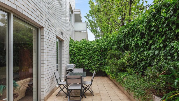 A patio with a table and chairs, a grill, and a hedge. - Lalique Elwood Beachside Apartment (Elwood)