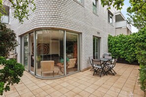 Eine sonnige Terrasse in der Ecke mit Glastür zu einem Innenraum, einem Esstisch mit Stühlen und einer grünen Hecke.