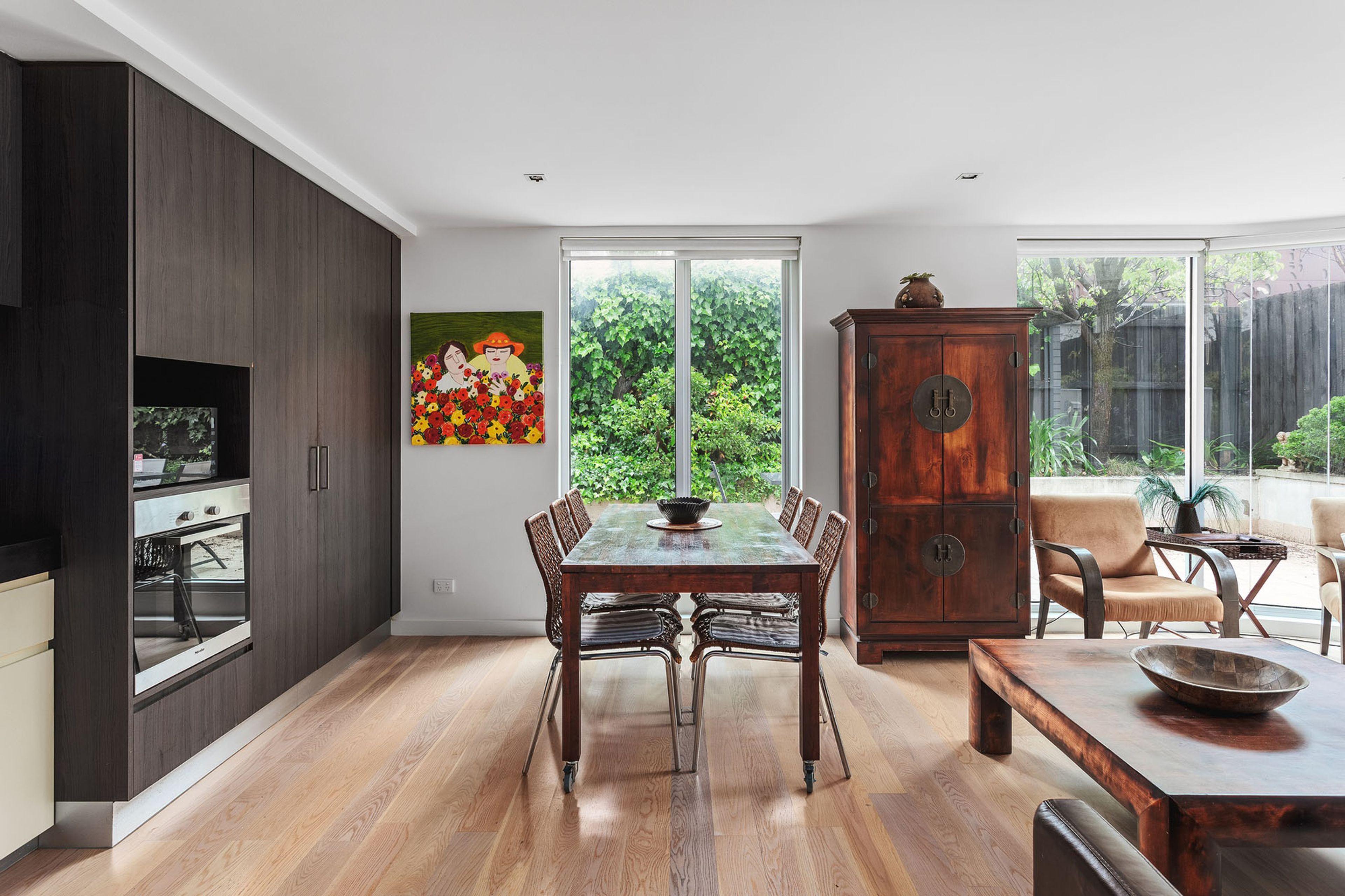 A modern kitchen with dark wood cabinets, a dining table with chairs, and a large window overlooking a garden.