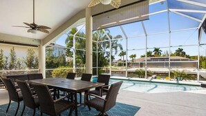 Ein Poolbereich mit Esstisch und Stühlen, Deckenventilator und großen Fenstern mit Blick auf einen Pool und Palmen.