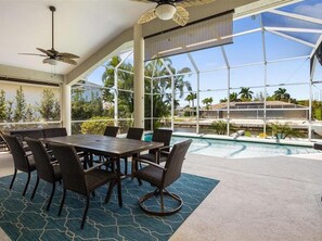 A poolside area with a dining table and chairs, a ceiling fan, and large windows overlooking a pool and palm trees.