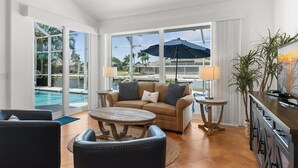 Ein geräumiges Wohnzimmer mit einer Couch, einem Couchtisch und Sesseln, großen Fenstern mit Blick auf einen Pool und Palmen, sowie eine Küchenzeile mit Bar.