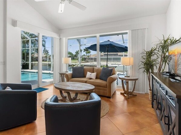 A spacious living room with a sofa, coffee table, and chairs, large windows with a view of a pool and palm trees, and a kitchen area with a bar.