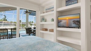 Ein Zimmer mit einem großen Bett, einem Fernseher auf einem weißen Regal, Blick auf einen Pool und einem Deckenventilator.