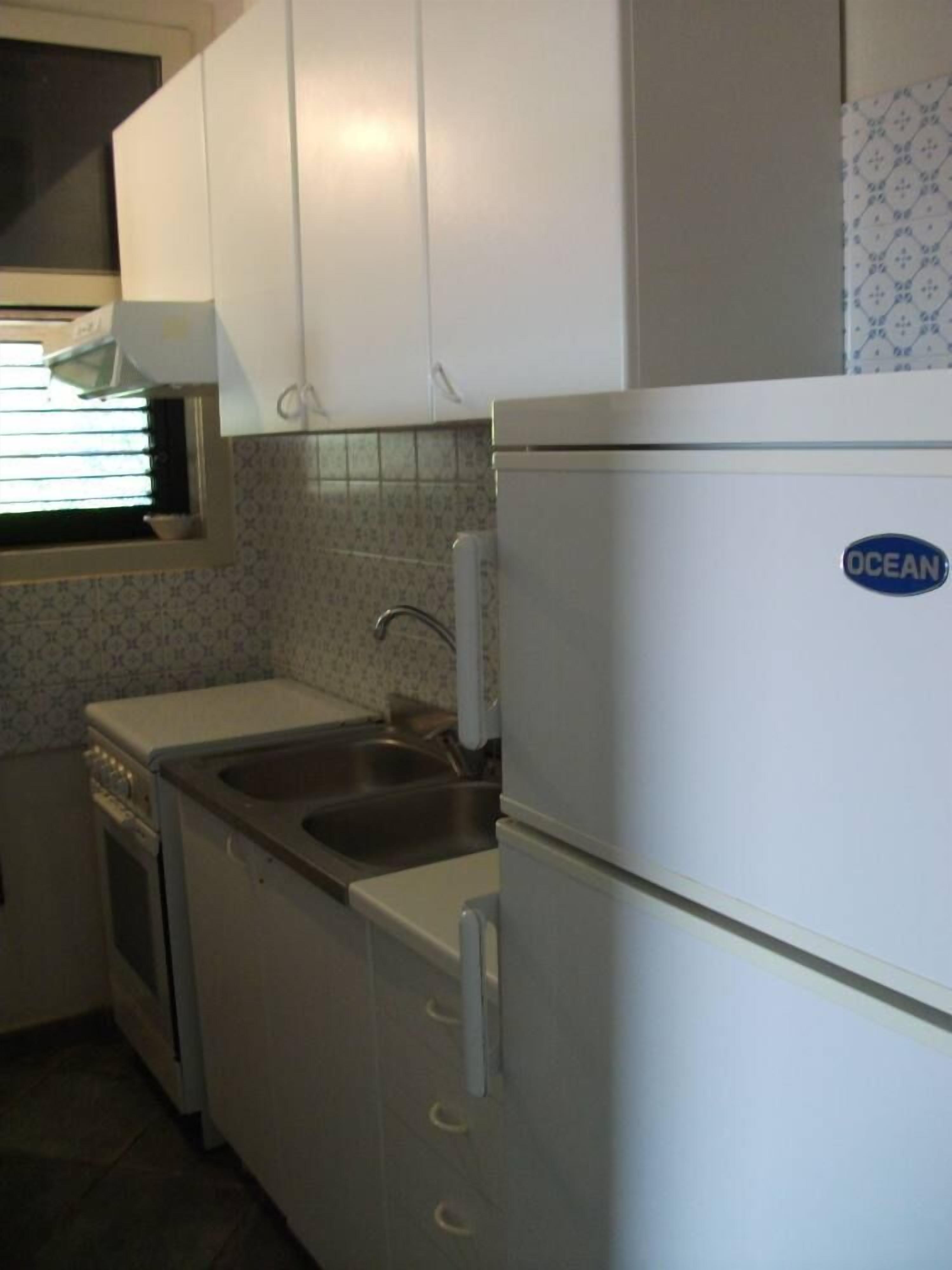 A compact kitchen with white cabinets, a stainless steel sink, and a white refrigerator with a blue logo.