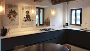 Une cuisine moderne avec des meubles de couleur sombre, une cuisinière à gaz et des luminaires suspendus. Deux fenêtres laissent entrer la lumière naturelle et une grande affiche orne le mur.