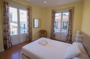 Ein Hotelzimmer mit einem großen Bett, zwei Fenstern mit floralen Vorhängen, einem Korbsessel und Blick auf Gebäude.