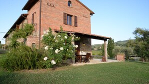 A rustic brick house with a covered patio, surrounded by greenery and a clear sky.