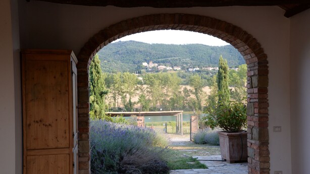 An arched doorway leading to a courtyard with a stone floor, surrounded by greenery and a view of a hillside village.