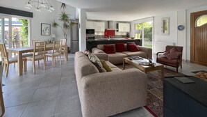Uma sala de estar espaçosa com sofá, mesa de centro e mesa de jantar com cadeiras. Uma cozinha visível ao fundo, com eletrodomésticos modernos.