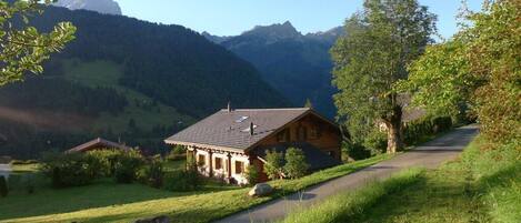 Ein hölzernes Haus mit geneigtem Dach, umgeben von üppiger Vegetation und einer Bergkulisse.