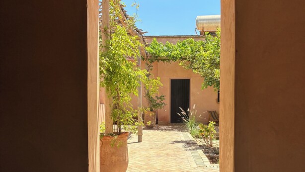 Une porte ouverte donne sur une cour intérieure, dont le sol est recouvert de carreaux, où l’on trouve des plantes en pot et un bâtiment doté d’une porte sombre.