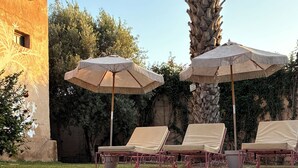 Un espace piscine doté de chaises longues et de parasols, entouré de verdure et bordé d’un bâtiment à la façade texturée.