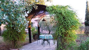 Un patio jardín con mesa y sillas, rodeado de follaje y una estructura de pérgola.