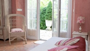 A bedroom with a bed, a wicker chair, a nightstand, and a door leading to a garden. - Country Cottage / Gite - LOUBERS (Loubers)