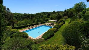 En rektangulär pool omgiven av välskötta häckar och träd, med en överbyggd sittplats och en klarblå himmel ovanför.