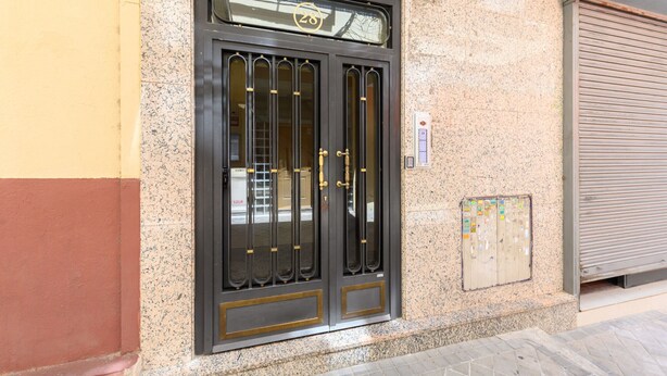 A building entrance with a large, ornate black door, a number plate above it, and a closed shutter to the right. - Downtown Madrid North Barrio del Pilar. Tempory (Madrid)