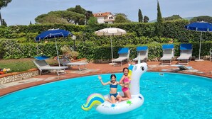 A swimming pool with lounge chairs and umbrellas, surrounded by greenery and a hedge. - L'ULIVETA DEL LUSCHI VILLA, PISCINA  E CAMPO DA TENNIS, WI-FI, PET FRIENDLY (ROSIGNANO MARITTIMO)