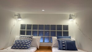 Una habitación pequeña y acogedora con ventana, dos camas y dos luces fijadas a la pared.