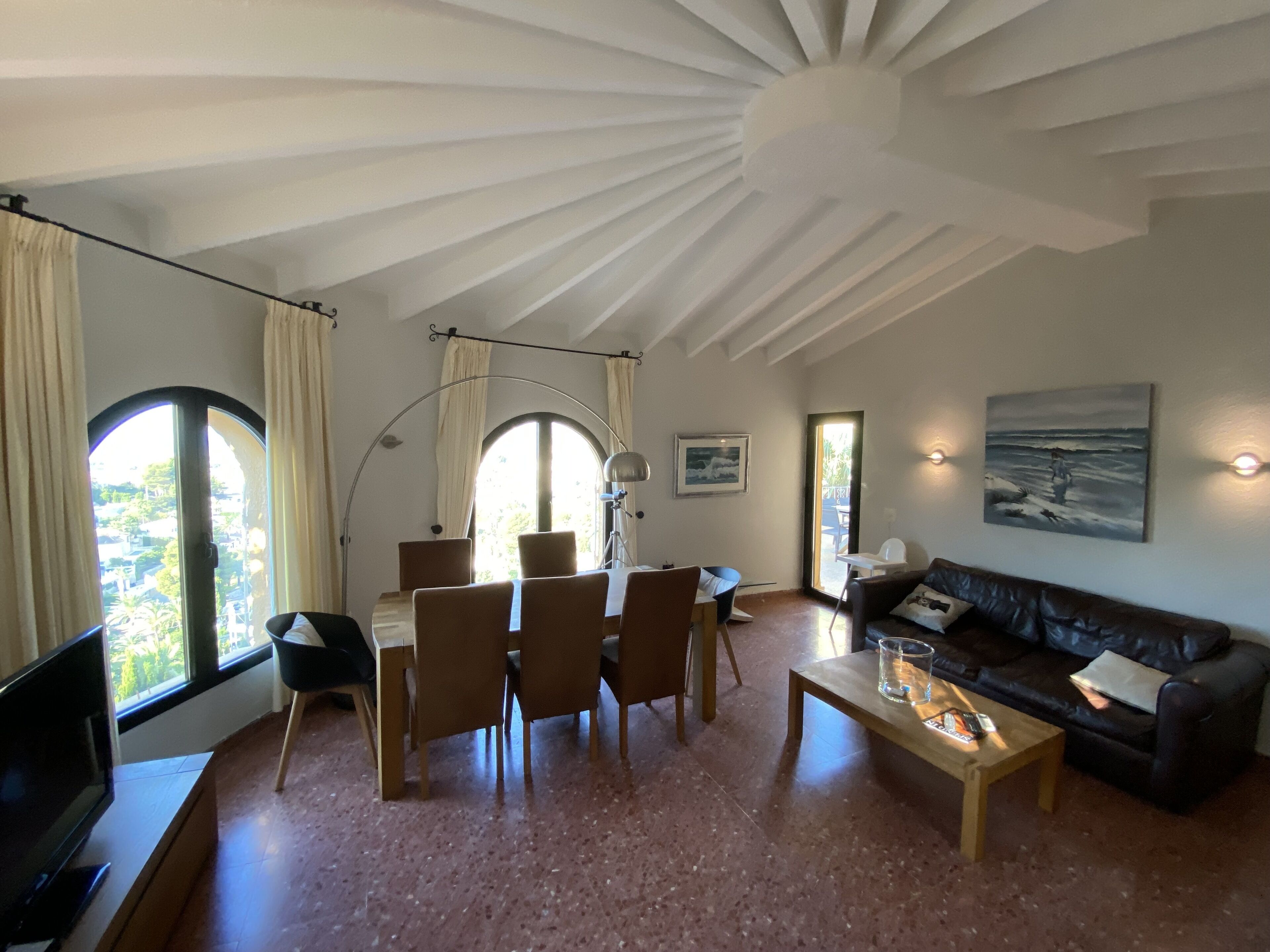A spacious living room with a high ceiling, a dining area with chairs and a table, a sofa, a flat-screen TV, and artwork on the walls.