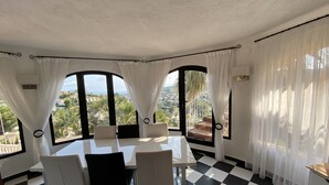 Un espace repas avec un sol à carreaux, une table et des chaises blanches, de grandes fenêtres avec des rideaux, et une vue sur la verdure et les bâtiments.