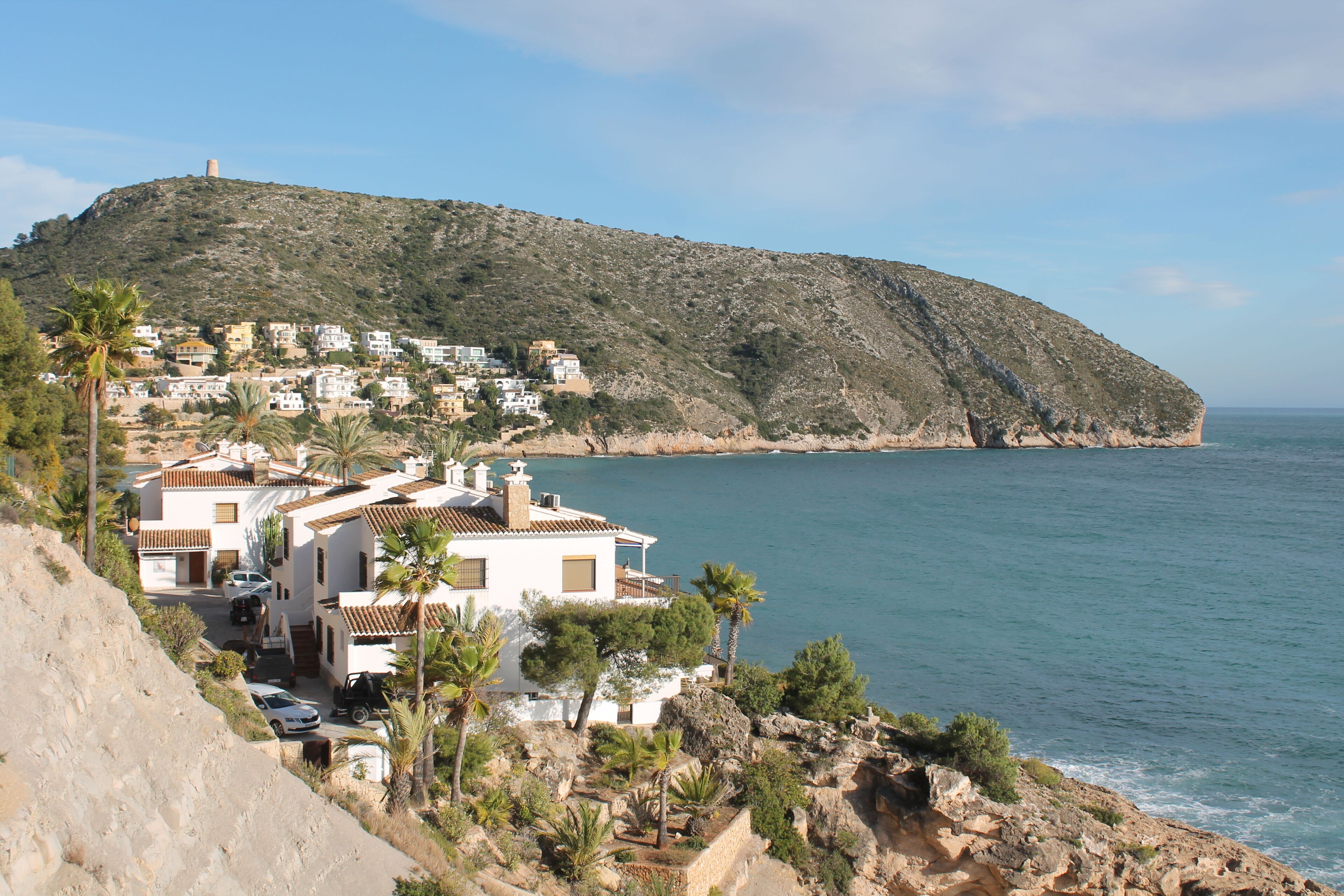A coastal town with white buildings, a hill with a tower, and a clear view of the sea.