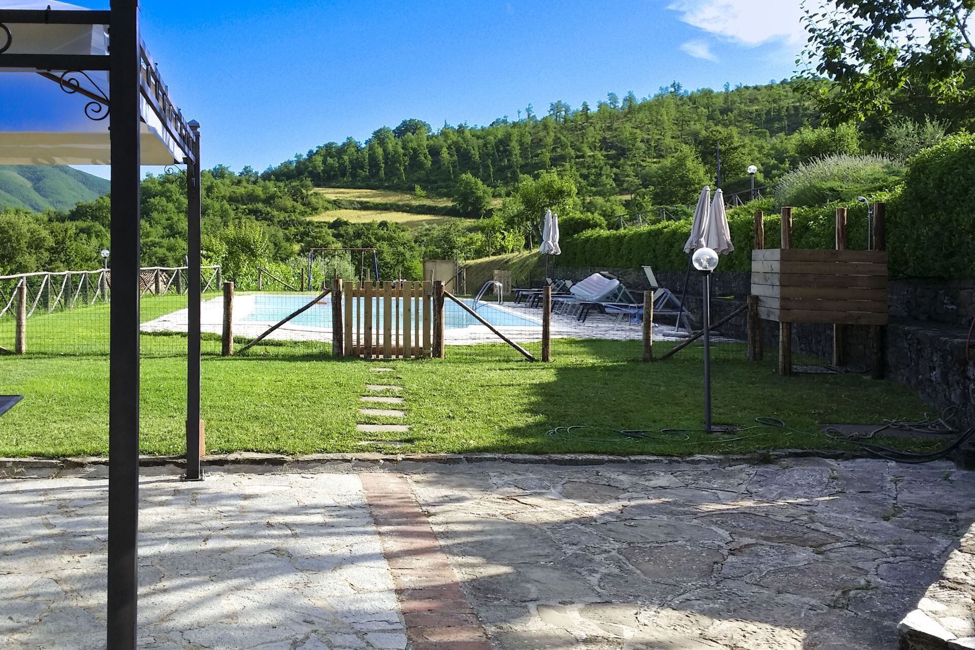 A pool area with a wooden fence, lounge chairs, and a pergola with umbrellas.