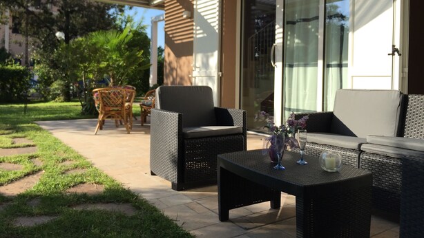 Eine Terrasse im Freien mit Korbmöbeln, einer Glastür zu einem überdachten Bereich und einem kleinen Tisch mit Kerze und Glas.