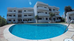 Une piscine à bord incurvé, entourée d’une terrasse en pierre. Au fond, on aperçoit un immeuble blanc à plusieurs étages, doté de balcons et de fenêtres.
