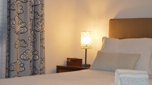Un lit bien fait, avec du linge de lit blanc, un coussin à rayures et une serviette pliée posée sur le côté. On aperçoit une table de chevet avec une lampe et une petite boîte en bois. Les rideaux sont ornés d’un motif floral.