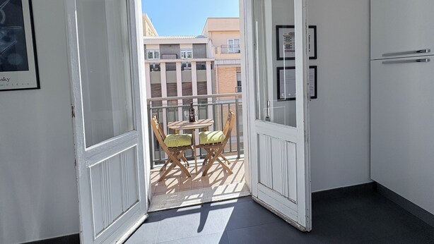 Un couloir menant à un balcon agrémenté d’une table et de chaises, un tableau encadré au mur et une porte avec une fenêtre.