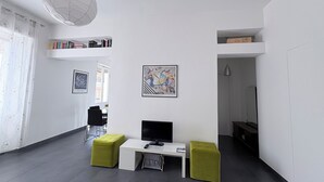 Un salon moderne avec un bureau blanc, deux poufs verts, un téléviseur à écran plat et des œuvres d’art accrochées aux murs.