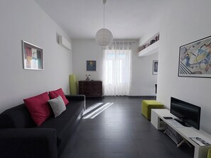A modern living room with a dark sofa, a flat-screen TV, a small table, and a bookshelf.