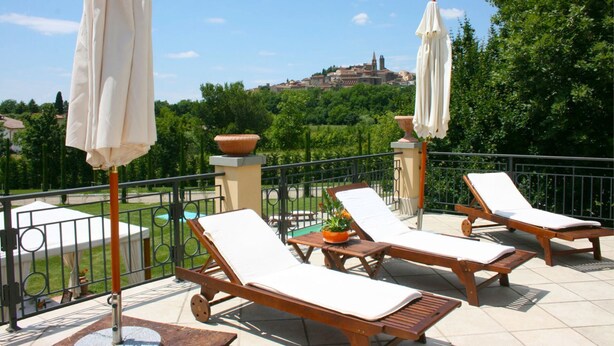 A terrace with lounge chairs and umbrellas, a view of a hill with a building, and a garden area.