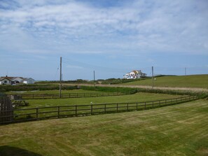 Paisaje rural con una casa en una colina, rodeada de campos cubiertos de hierba y una valla de madera.