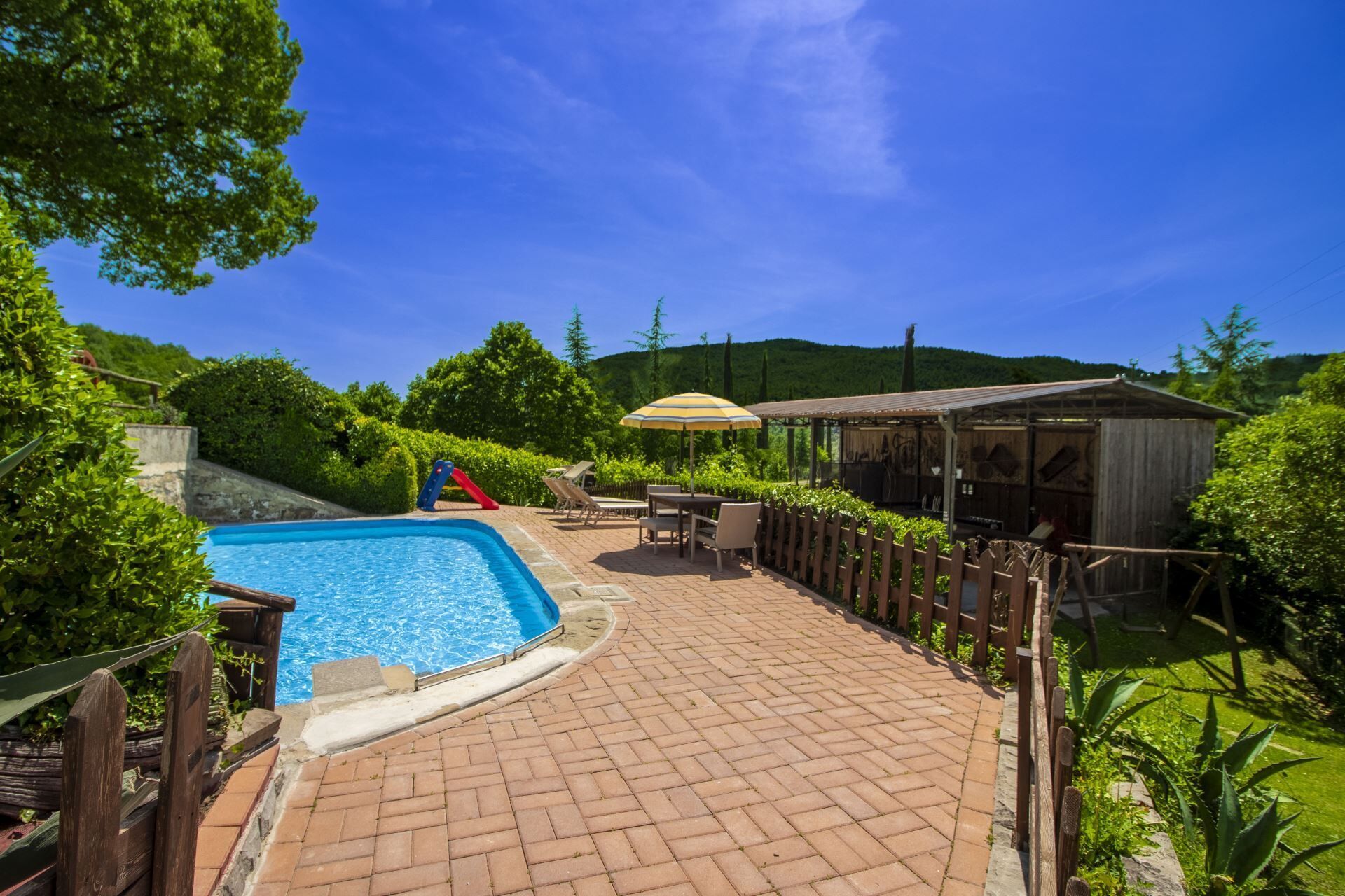 A swimming pool with a slide, a wooden deck, and a covered area with tables and chairs.